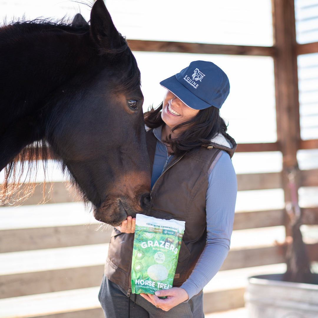 Woman holding a package of horse treats next to a horse in a stable.