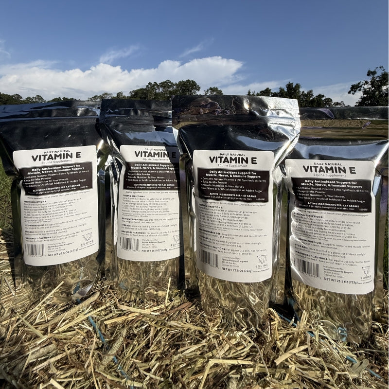 Four stand-up pouches of  Natural Vitamin E shown outdoors on a hay bale, with a blue sky and trees in the background.