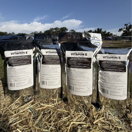 Four stand-up pouches of  Natural Vitamin E shown outdoors on a hay bale, with a blue sky and trees in the background.
