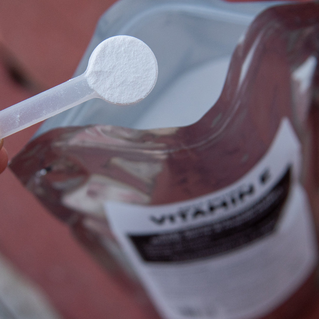 Hand holding a spoon with a vitamin supplement over a pouch labeled &
